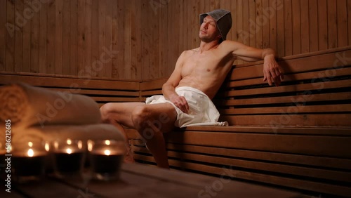 Wide shot sweating Caucasian man sitting in wooden Finn sauna with burning candles. Portrait of confident guy enjoying leisure indoors in SPA. Relaxation and tranquility concept