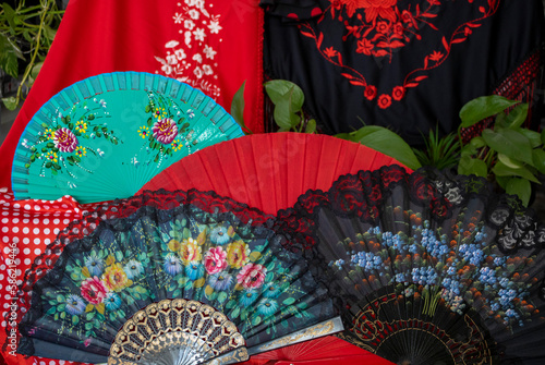 Close-up fans of different colors, red and black with flowers and lace, green fan with flowers and shawls in the background, one black and one red, typical Andalusian topos. April fair