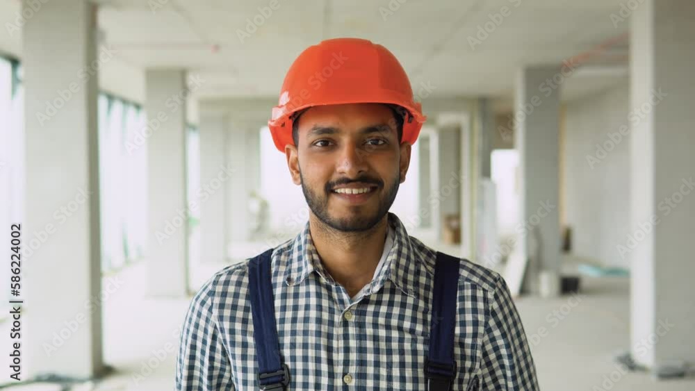 Portrait of Indian worker builder in uniform wearing a safety helmet looking at the camera, standing on construction site Portrait of Indian worker builder in uniform wearing a safety helmet looking at the camera, standing on construction site