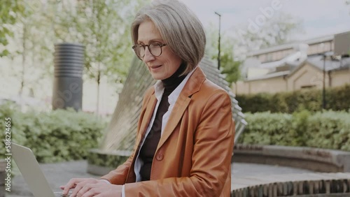 Senior woman with glasses and casual stylish clothes working on laptop, typing message, browsing Internet while sitting on park bench outdoors. Elderly woman freelancer, active retired working outside