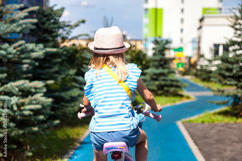 Rear view of little child in pink hat riding bike in summer park, outdoors. Little blonde girl kid enjoy riding small bicycle outside, summertime. Childhood leisure activity concept. Copy text space