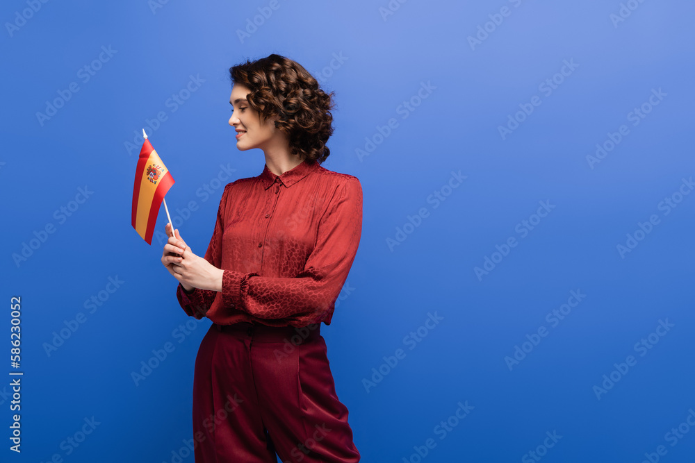 happy language teacher with curly hair holding flag of Spain isolated on blue.