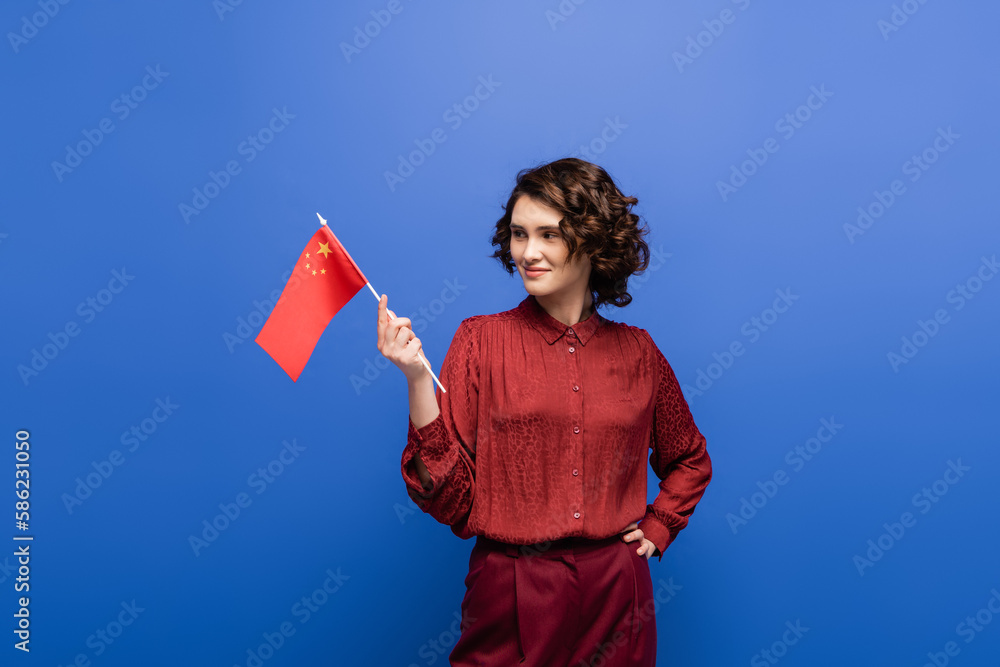 happy language teacher holding flag of China while standing with hand on hip isolated on blue. happy language teacher holding flag of China while standing with hand on hip isolated on blue.