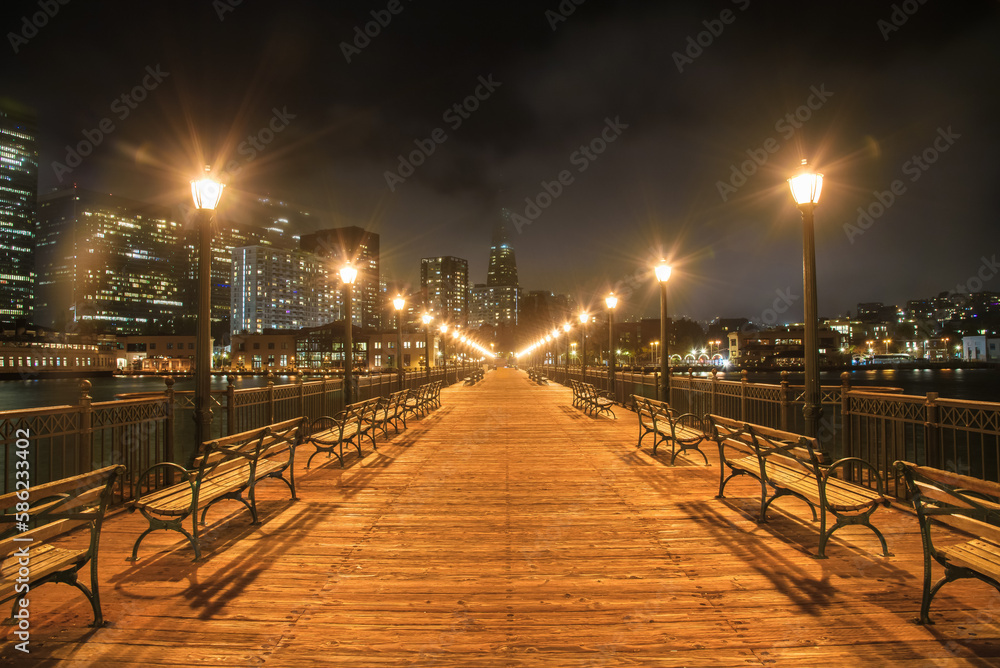 Deserted Pier seven  lined with street lights and benches in San Francisco at night. Downtown skyline partly covered with fog is in background.