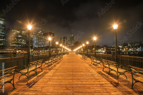 Deserted Pier seven lined with street lights and benches in San Francisco at night. Downtown skyline partly covered with fog is in background.