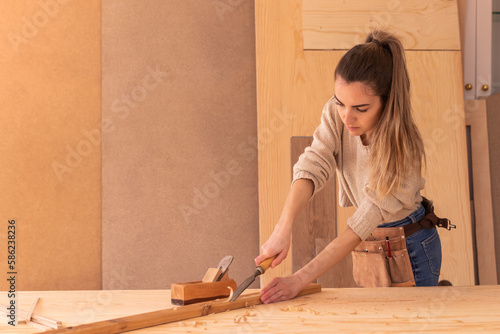 Craftswoman carving wood with chisel. Concentrated female joiner in apron gouging wood with chisel near jack plane photo