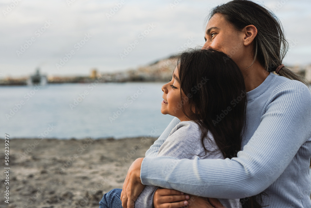 Happy latin mother hugging her little daughter on the beach while looking the sunrise together - Family and love concept