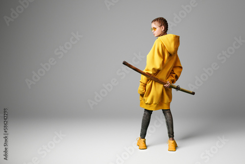 A girl in a yellow hoodie with a large Japanese sword in a wooden scabbard on a gray background. Studio portrait