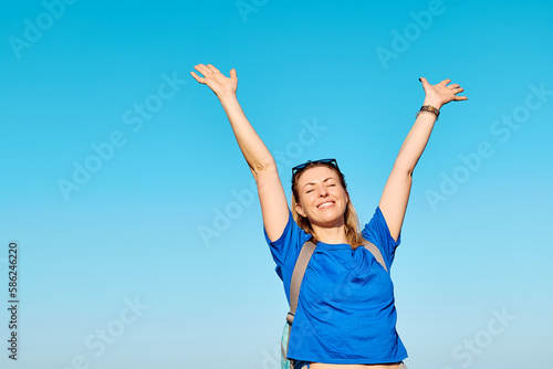 Blond woman tourist with closed eyes spreading arms up to sky enjoying freedom by the sea. Female traveler relaxing in serene nature. Mental health, wellness, adventure travel and healthy lifestyle.