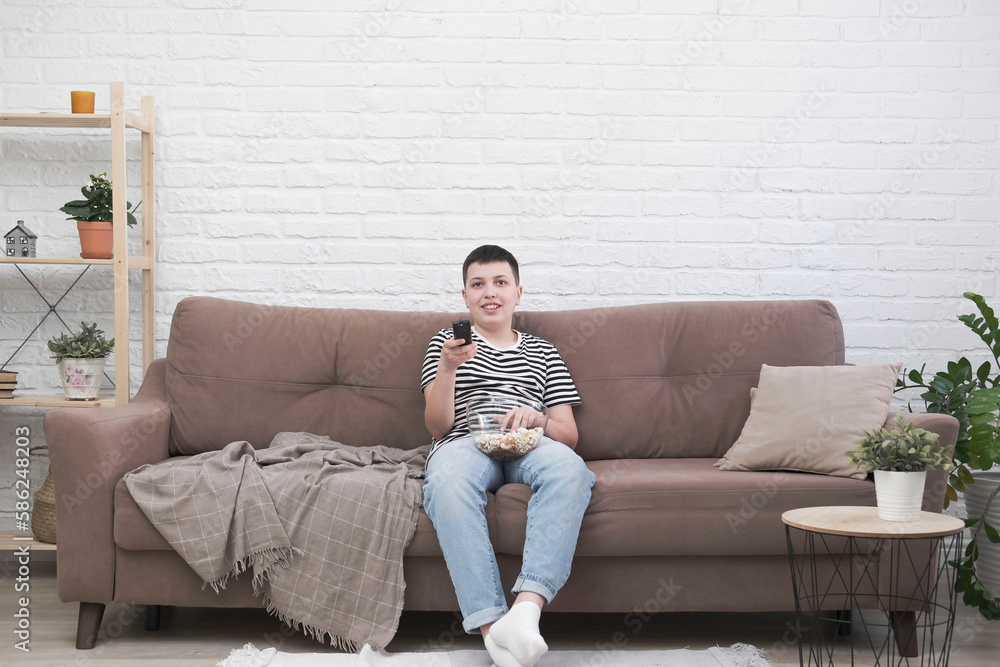 Obraz premium A teen boy is sitting on the sofa, eating pop corn, holding control panel and watching tv in light living room.