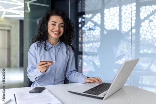 Portrait of a young Muslim business woman sitting in the office at the table with a laptop. Holds a credit card, works with accounts, online shopping. She looks at the camera smiling.