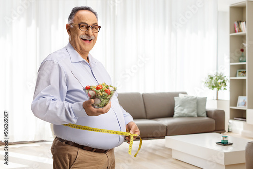 Mature man holding a salad and measuring waist in a living room