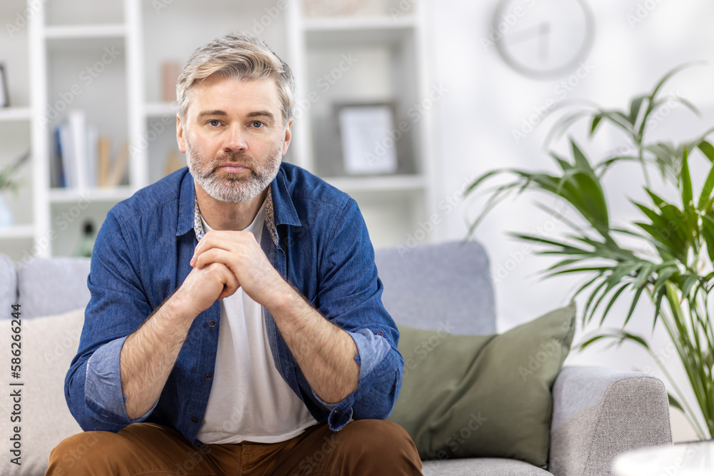 Portrait of a scary handsome gray-haired man in a blue shirt sitting on the sofa at home and seriously looking at the camera, crossing his arms in front of him. Portrait of a scary handsome gray-haired man in a blue shirt sitting on the sofa at home and seriously looking at the camera, crossing his arms in front of him.