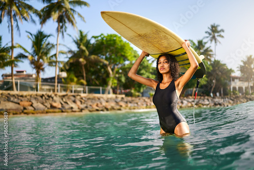 African american woman standing with surfboard on head in ocean water. Black female surfer posing with surf board. Pretty multiethnic girl goes from surfing session on tropical location.