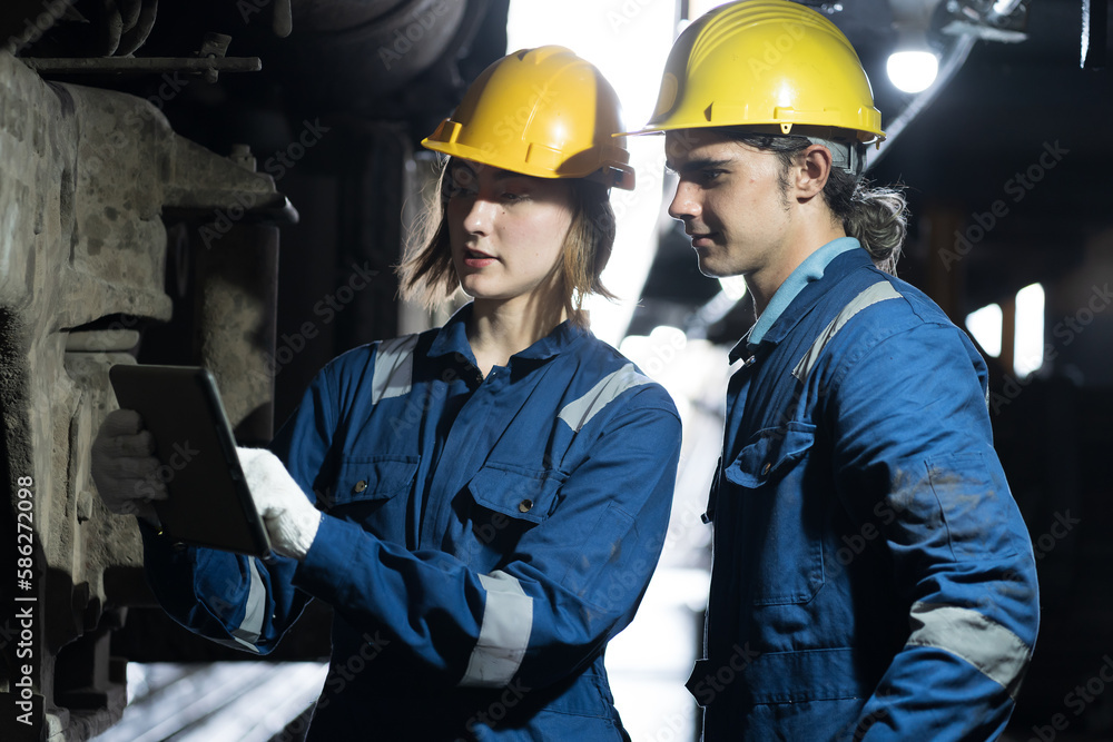 Male and female engineer maintenance locomotive engine, wearing safety uniform, helmet and gloves in locomotive garage. Male and female railway engineer use tablet while repair train wheel in garage Male and female engineer maintenance locomotive engine, wearing safety uniform, helmet and gloves in locomotive garage. Male and female railway engineer use tablet while repair train wheel in garage