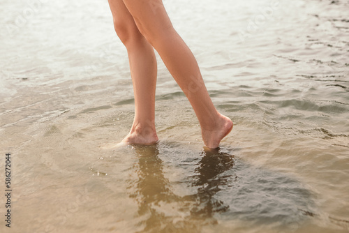 Close up naked slim woman legs walking barefoot along the river. Female feet on sea beach with splashing waves and foam. Girl tourist on vacation. Lady go on shore after bathing 