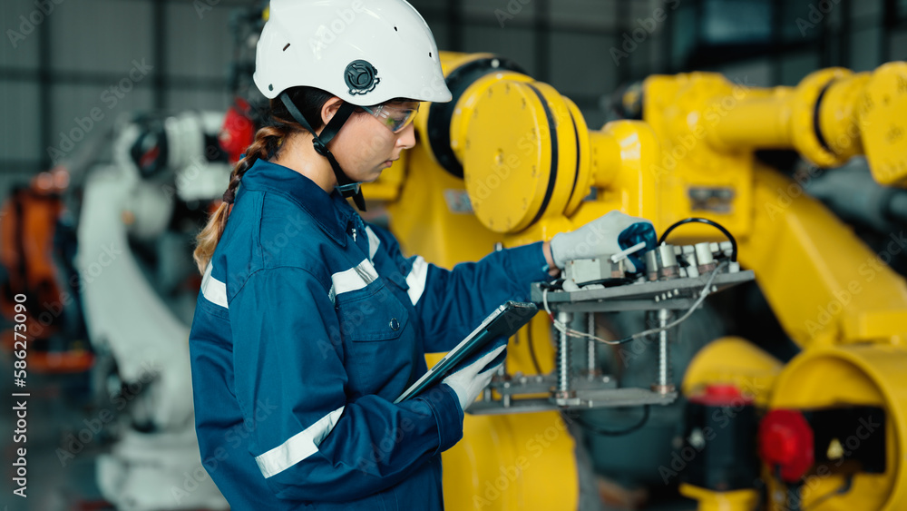 Caucasian mechatronics engineer is using a tablet to record the results of examining a lot of robotic arms. A female industrial worker with responsibility for quality checking machines in the factory. Caucasian mechatronics engineer is using a tablet to record the results of examining a lot of robotic arms. A female industrial worker with responsibility for quality checking machines in the factory.