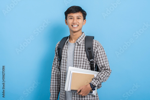 Young asian college student carrying books and backpack isolated over blue background