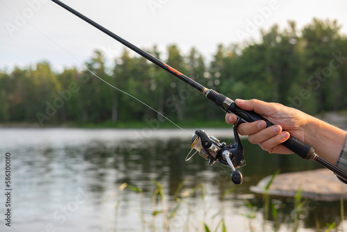 Close up of a man's hand holding a fishing rod. Sunset soft light, selective focus, blurred forest and lake in the background. Vacation, camping, fishing, relaxing and hobby concept.