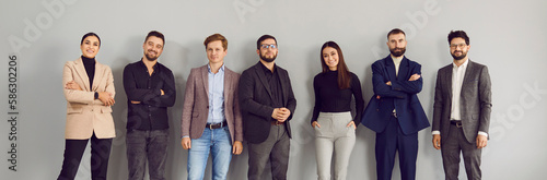 Happy smiling professional young business people, attractive men and women lined up along the wall, confidently looking at the camera. Panoramic shot on isolated gray background.