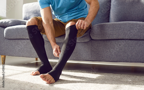 Cropped photo of a man putting on black medical compression stockings on his legs for the prevention of varicose veins and for venouse therapy sitting on the couch at home. Healthcare concept. photo