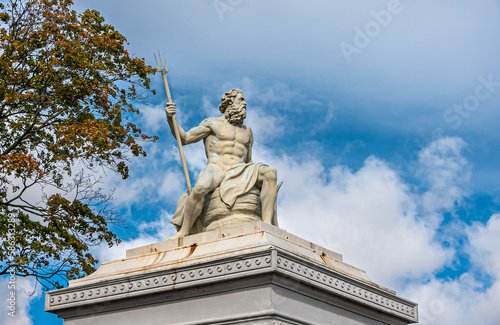 Copenhagen  Denmark - September 13  2010  Closeup  Neptunus or Poseidon God white stone statue on pillar at east side entrance to Churchill Parken. Blue cloudscape and green foliage