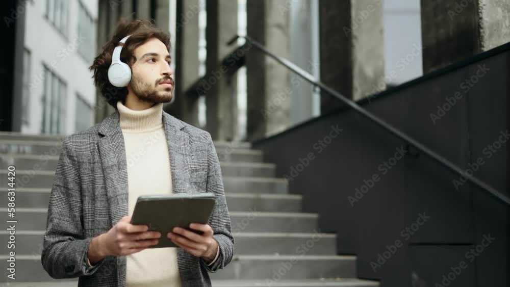 Handsome young arabian businessman with headphones working on tablet gadget near modern business centre Busy muslim man have distance remote freelance work outdoors Handsome young arabian businessman with headphones working on tablet gadget near modern business centre Busy muslim man have distance remote freelance work outdoors