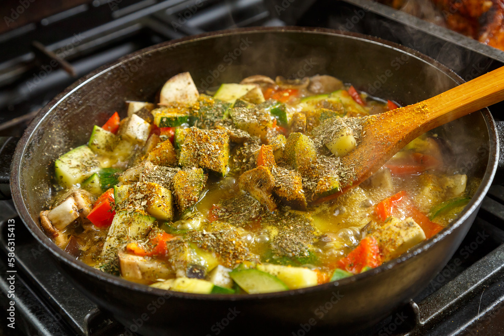 A frying pan with stewed vegetables and a wooden spoon on the stove . A frying pan with stewed vegetables and a wooden spoon on the stove .