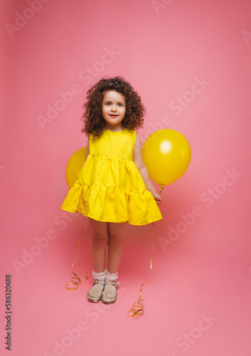 Portrait of a cheerful little girl isolated on a pink background, holding a bunch of colorful balloons, posing.