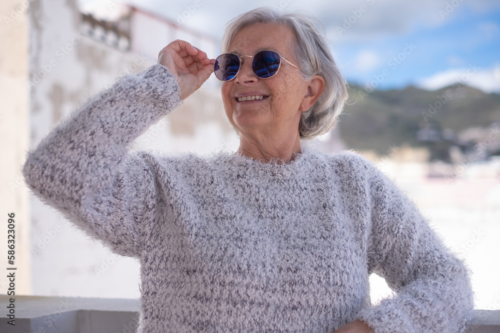 Portrait of happy senior woman smiling holding her glasses. Face of attractive senior woman wearing blue reflective sunglasses outdoors. Relaxed retired woman with gray hair