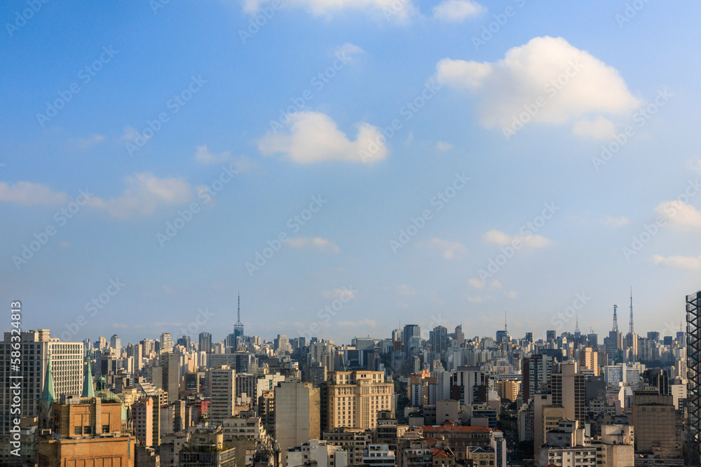 Cidade de São Paulo vista do alto, com edifícios, torres antenas e céu azul com nuvens.