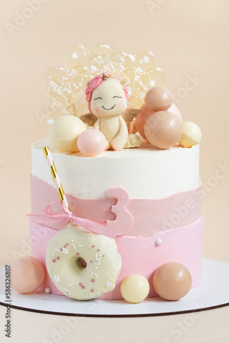 Birthday cake for a girl with pink pastel cream cheese frosting decorated with unicorn figure, chocolate spheres and caramel candies on the beige background. © alexanderon