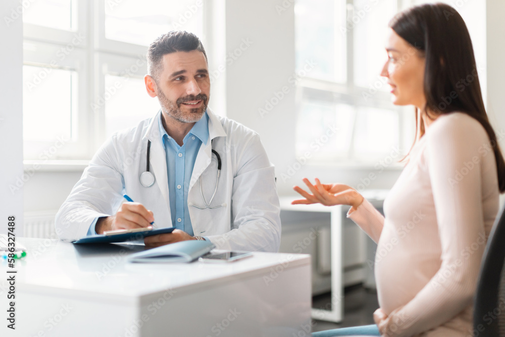 Young pregnant woman sitting at hospital, talking and having consultation with middle aged male gynecologist Young pregnant woman sitting at hospital, talking and having consultation with middle aged male gynecologist