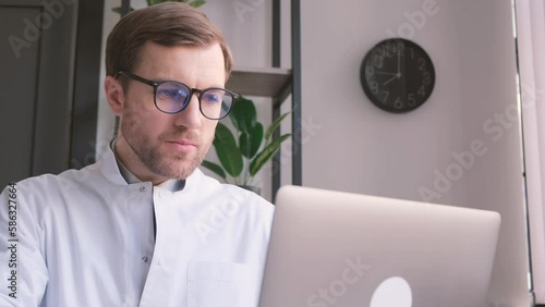 Focused serious young male doctor in glasses and white uniform working on computer. photo