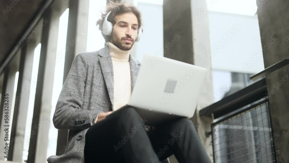 Handsome young arabian businessman with headphones working on laptop sitting near modern business centre Busy focused muslim man have distance remote freelance work on computer outdoors alone Handsome young arabian businessman with headphones working on laptop sitting near modern business centre Busy focused muslim man have distance remote freelance work on computer outdoors alone