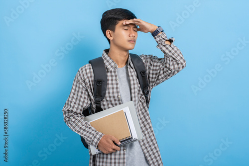 Young asian college student carrying books and backpack feeling sick tired and frustration isolated over blue background photo