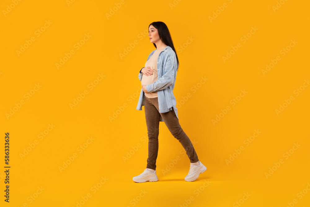 Young pregnant woman in casual wear, walking and touching her belly on yellow studio background, full length, free space Young pregnant woman in casual wear, walking and touching her belly on yellow studio background, full length, free space