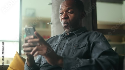 Portrait of mature smiling african american man looking at smartphone cellphone screen with nod of approval sitting on sofa at home and giving thumbs up Advise Approve Recommend Choice concept