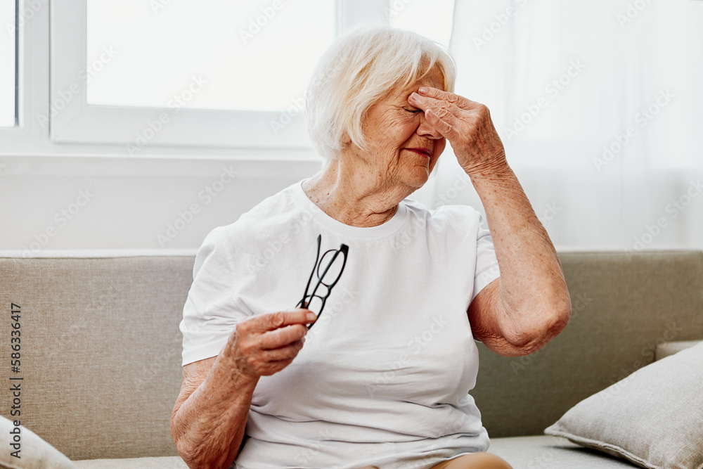 Elderly woman severe eye pain sitting on the couch, health problems in old age, poor quality of life. A grandmother with gray hair holds on to her sore eyes with glasses in her hand. Elderly woman severe eye pain sitting on the couch, health problems in old age, poor quality of life. A grandmother with gray hair holds on to her sore eyes with glasses in her hand.