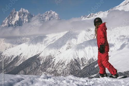 Young woman snowboarder wearing red ski suit and helmet riding on a slope against backdrop of stunning mountains in ski resort