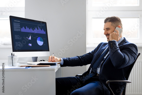 Confident middle aged businessman working on computer with graphics and charts and having business call in office