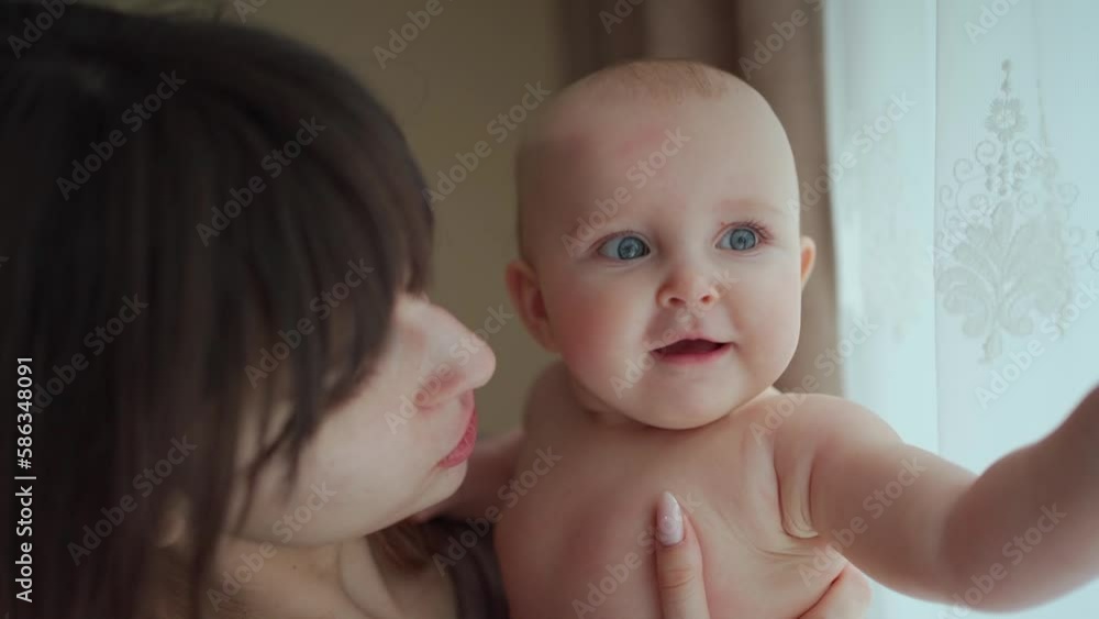A young charming mother holds her cute baby in her arms, gently kisses her. The child looks out the window, smiles and plays with tulle. Mother's Day A young charming mother holds her cute baby in her arms, gently kisses her. The child looks out the window, smiles and plays with tulle. Mother's Day