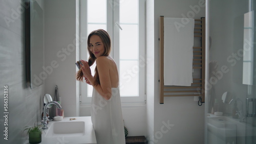 Positive lady arranging hair bathroom interior. Smiling woman holding hairbrush
