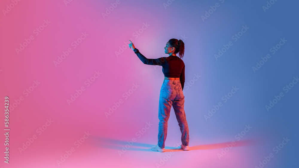 Back view of black lady touching imaginary screen, using virtual interface on colorful neon background, panorama