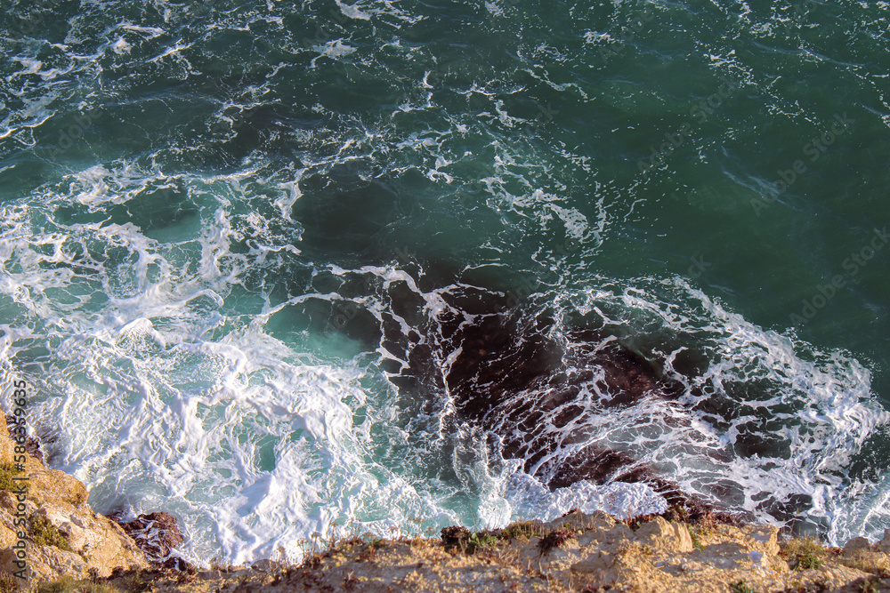 Dark turquoise waves breaking on black rocks, rolling tide, bird's eye view. Ocean photography. 