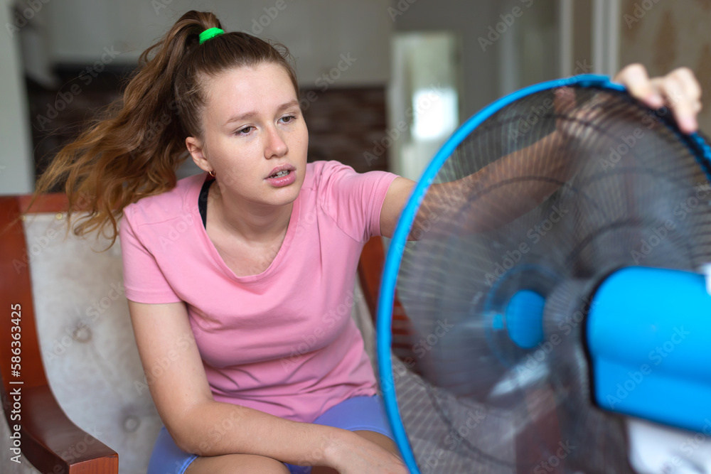 Young woman using electric fan at home in living room, sitting on couch