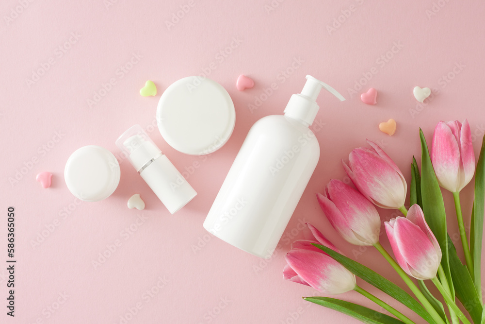 Natural cosmetic products concept. Top view photo of pump bottle without label cream jars colorful hearts and bouquet of pink tulips on pastel pink background