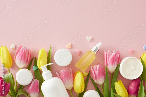 Organic skincare concept. Top view photo of dropper bottle pump bottle without label cream jars colorful hearts and pink yellow tulips on isolated pastel pink background with copyspace