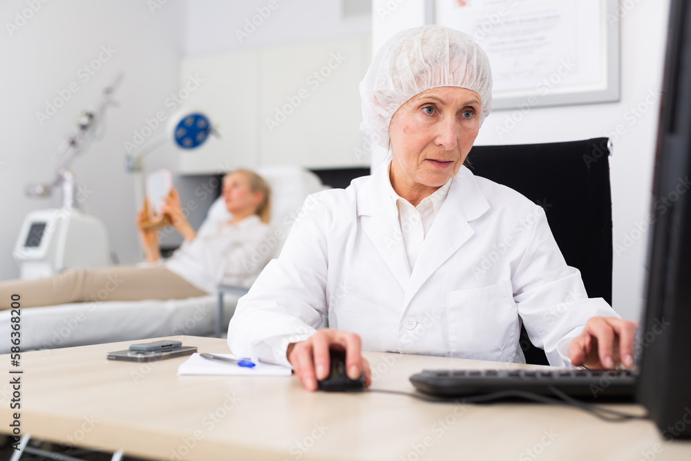 Senior woman dermatologist using computer during appointment. Her patient waiting in background.