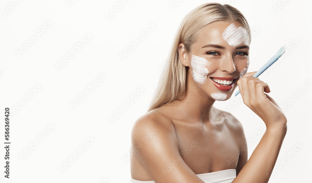 Smiling blond girl uses scrub, skincare cream with brush. Portrait of young woman washing face, using her cosmetic products for better skin tone, standing over white background Smiling blond girl uses scrub, skincare cream with brush. Portrait of young woman washing face, using her cosmetic products for better skin tone, standing over white background
